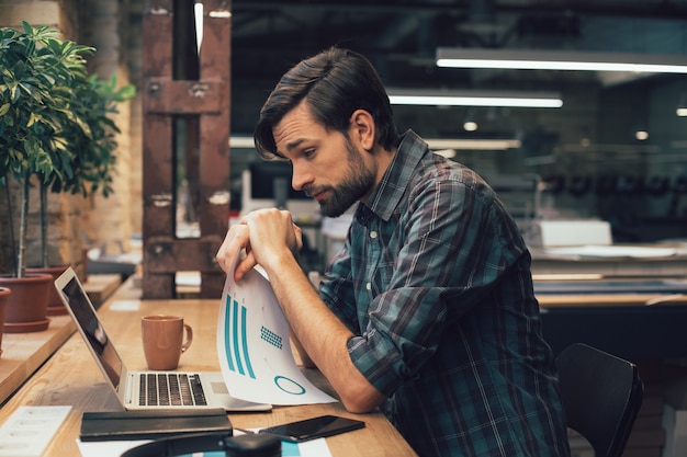 Homem cansado segurando uma folha de papel com um diagrama e olhando para a tela do laptop