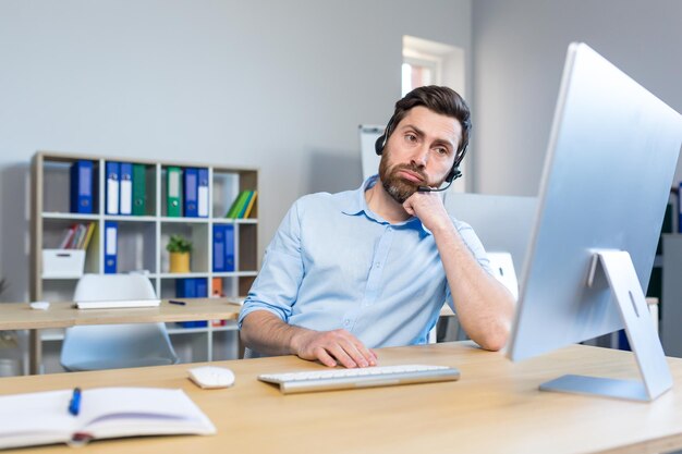 Homem cansado trabalhando em um computador com um fone de ouvido para uma chamada de vídeo no escritório