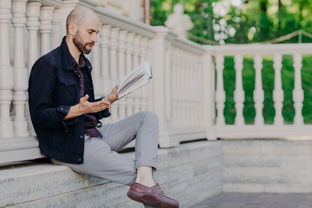 Homem careca atraente lê atentamente gestos de livros interessantes com a mão tem um enredo impressionante tenta entender a sensação das poses dos personagens principais ao ar livre desfruta de ar fresco e vista maravilhosa