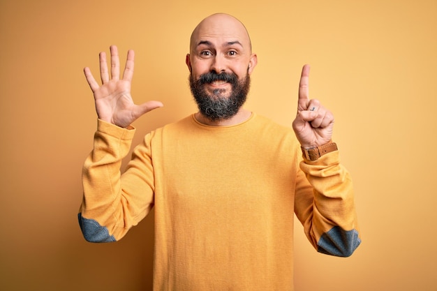 Homem careca bonito com barba vestindo suéter casual em pé sobre fundo amarelo mostrando e apontando para cima com os dedos número seis enquanto sorria confiante e feliz