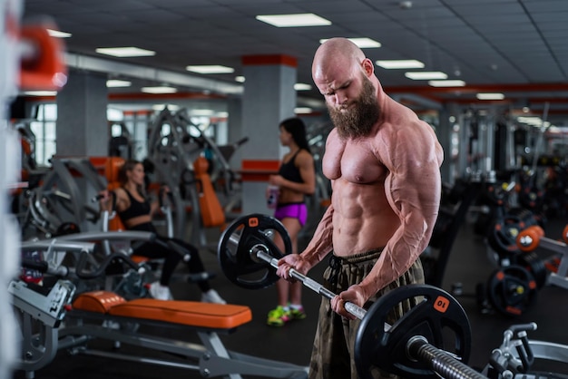 Homem careca com barba no ginásio Fisiculturista musculoso fazendo exercícios com barra Homem forte com torso nu O jovem atleta está se preparando para competições de levantamento de peso