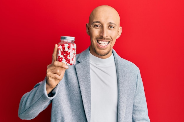 Homem careca com barba segurando um pote cheio de pílulas parecendo positivo e feliz em pé e sorrindo com um sorriso confiante mostrando os dentes