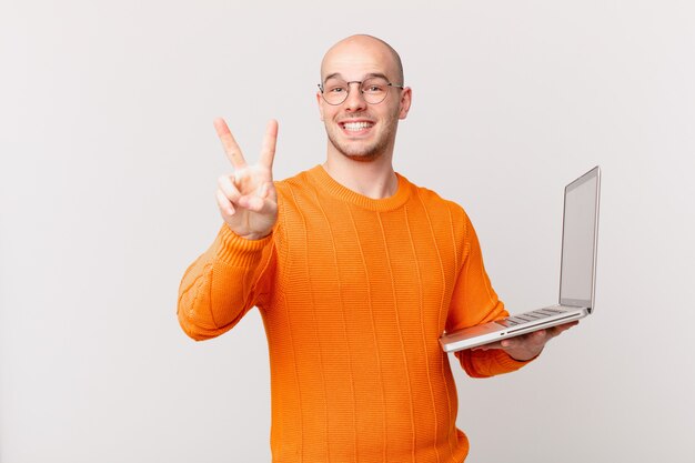 Homem careca com computador sorrindo e parecendo feliz, despreocupado e positivo, gesticulando vitória ou paz com uma mão