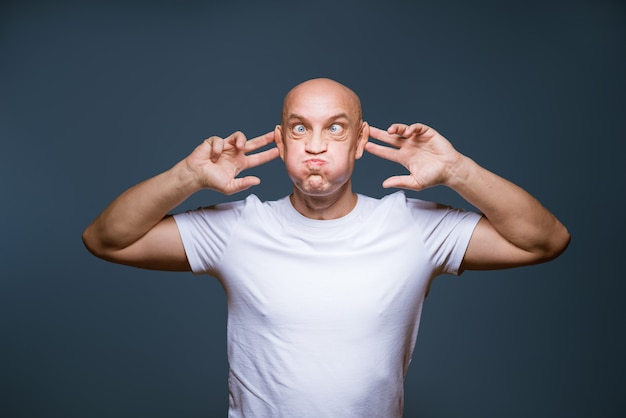 Homem careca cômico posando em uma parede azul com expressões faciais animadas e mãos levantadas na cabeça