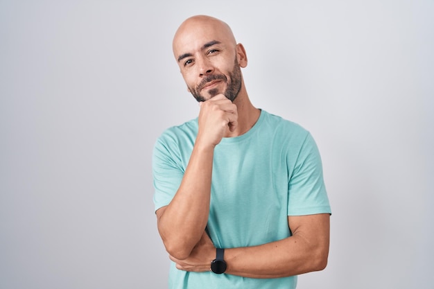 Homem careca de meia-idade em pé sobre fundo branco olhando confiante para a câmera sorrindo com braços cruzados e mão levantada no queixo pensando positivo