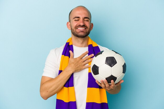 Homem careca do esporte jovem vestindo um lenço isolado no fundo azul ri alto, mantendo a mão no peito.