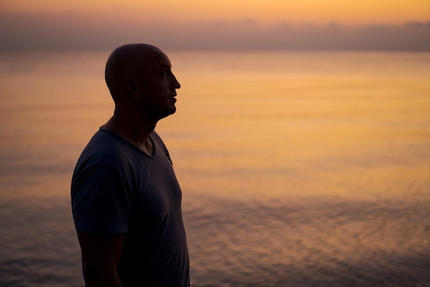 Homem careca parado na praia e olhando o incrível pôr do sol e mar com calma