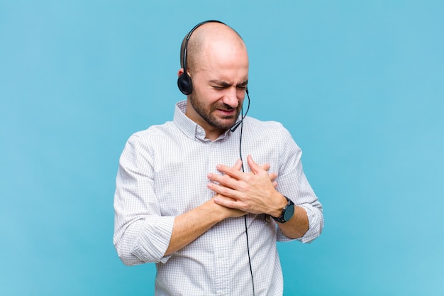 Homem careca parecendo triste, magoado e com o coração partido, segurando as duas mãos perto do coração, chorando e se sentindo deprimido