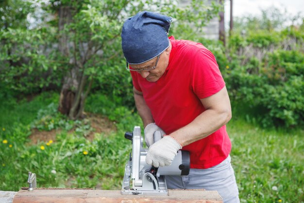 Homem carpinteiro com luvas serra com uma serra elétrica circular. Camiseta vermelha, calça cinza, no contexto da grama verde e árvores. Trabalho manual, construção de casas, ferramentas.