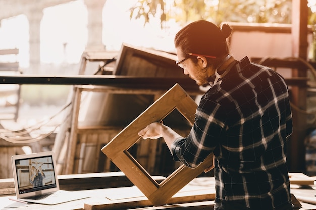 Foto homem carpinteiro que trabalha em uma oficina de bricolage aprende com a internet. autoeducação a partir do estudo de e-learning pela internet fazendo móveis a partir de vídeo em um laptop durante o trabalho de confinamento em casa.