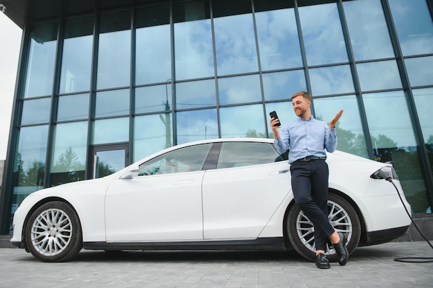 Foto homem carregando seu carro elétrico na estação de carga e usando smartphone