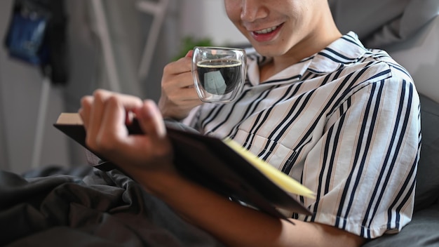 Homem casual bebendo café quente e lendo um livro na cama pela manhã