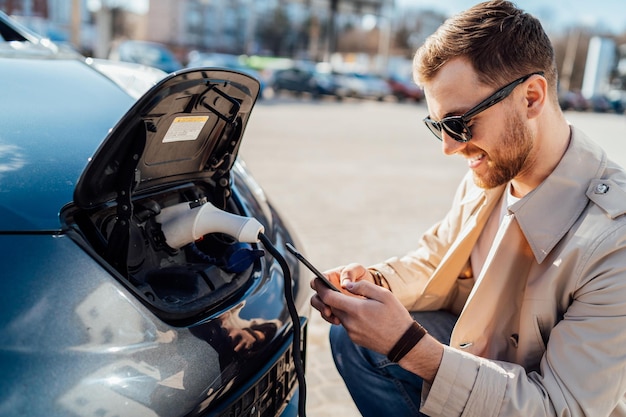 Homem casual com smartphone perto de carro elétrico