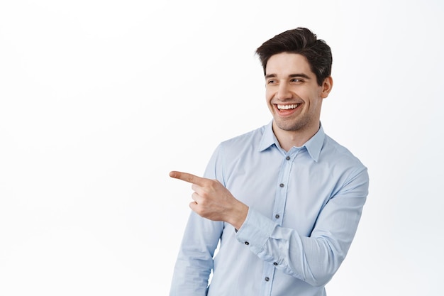 Homem caucasiano alegre com camisa do escritório, apontando e olhando para a esquerda com um sorriso satisfeito, mostrando algo legal, elogiando algo incrível, em pé sobre um fundo branco