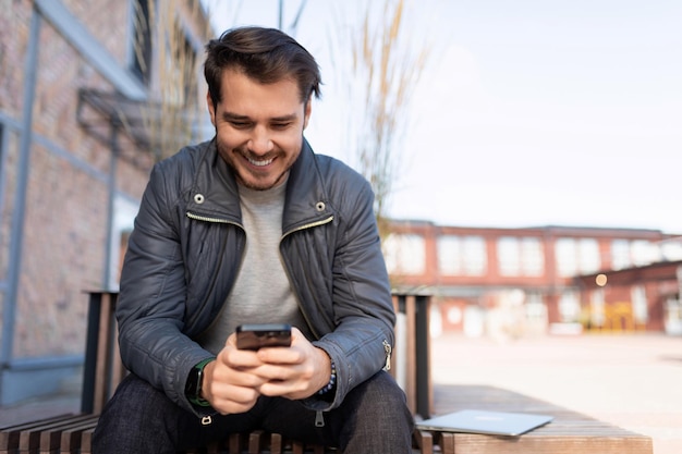 Homem caucasiano alegremente com um sorriso olha para a tela de um smartphone enquanto está sentado em um parque da cidade