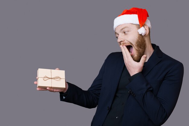 Homem caucasiano animado com barba segurando um presente e cobrindo a boca, gesticulando felicidade na parede cinza com espaço livre
