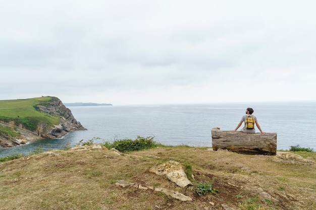 Homem caucasiano aventureiro isolado sentar em um banco do penhasco. Vista panorâmica horizontal do homem com a mochila viajando na costa. Conceito de pessoas e viagens.