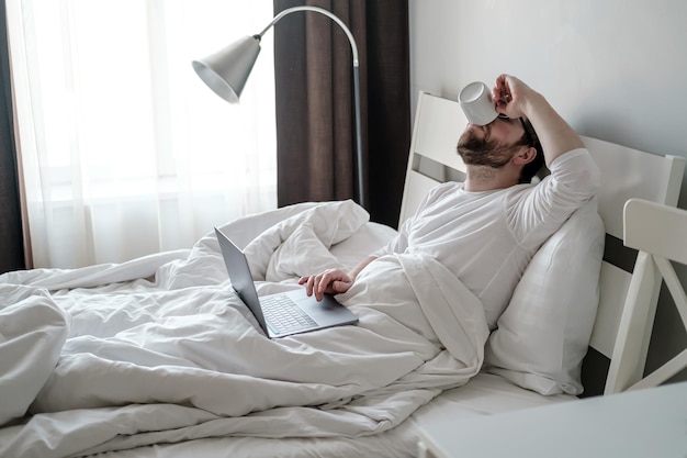 Homem caucasiano bebe café enquanto está sentado na cama de pijama ele faz uma pausa enquanto trabalha remotamente