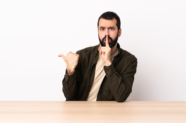 Homem caucasiano com barba em uma mesa apontando para o lado e fazendo gesto de silêncio