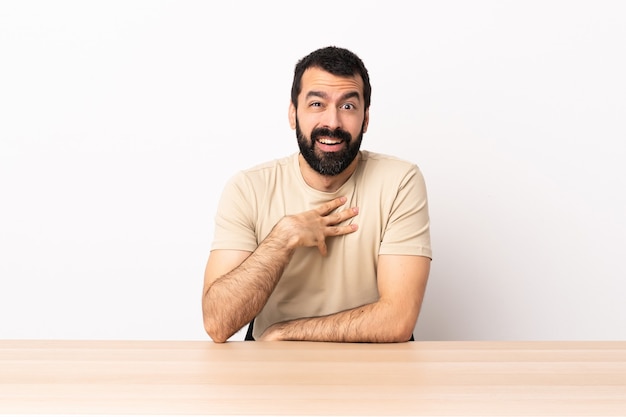 Homem caucasiano com barba em uma mesa apontando para si mesmo.