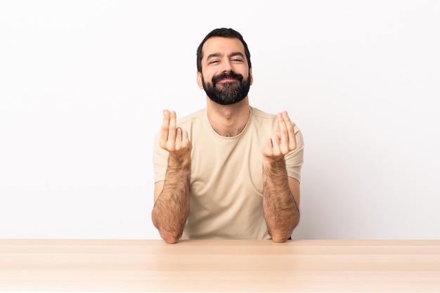 Homem caucasiano com barba em uma mesa fazendo gesto de dinheiro.