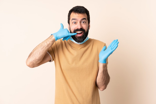 Homem caucasiano com barba, protegendo com uma máscara e luvas por cima da parede, fazendo o gesto do telefone e duvidando