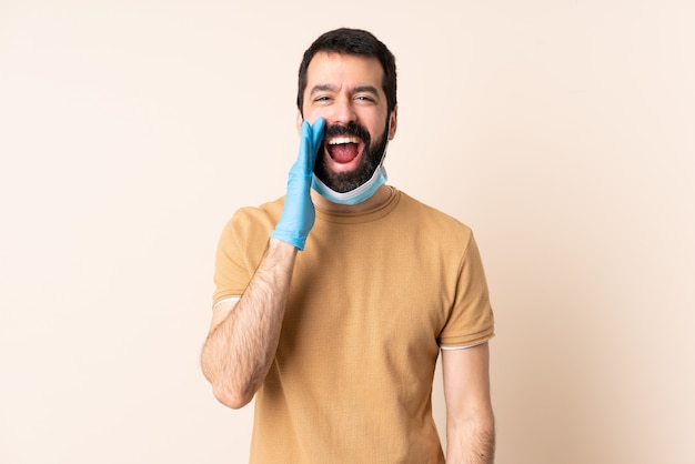 Homem caucasiano com barba, protegendo com uma máscara e luvas sobre parede isolada, gritando com a boca aberta