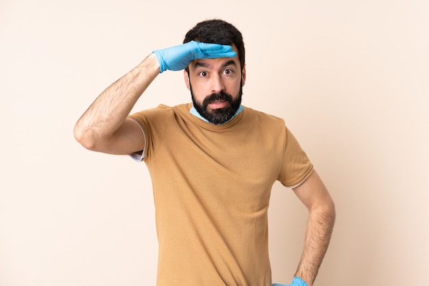 Homem caucasiano com barba, protegendo com uma máscara e luvas sobre parede isolada, olhando longe com a mão para procurar algo