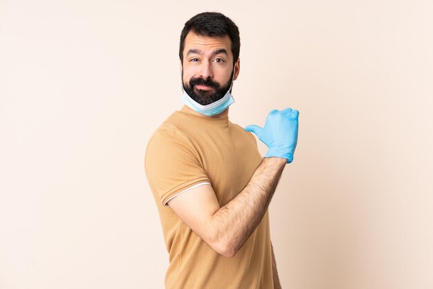 Homem caucasiano com barba, protegendo com uma máscara e luvas sobre parede orgulhoso e satisfeito