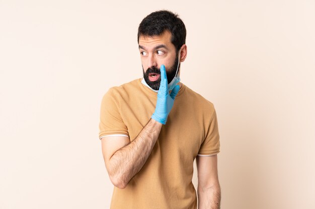 Homem caucasiano com barba, protegendo-se do coronavírus com uma máscara e luvas sobre a parede sussurrando algo com gesto surpreso enquanto olha para o lado