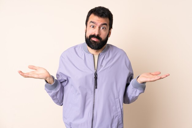 Homem caucasiano com barba, vestindo uma jaqueta sobre uma parede isolada, tendo dúvidas ao levantar as mãos