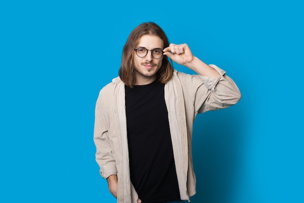 Homem caucasiano com cabelo comprido e barba tocando os óculos, sorrindo para a câmera na parede azul do estúdio