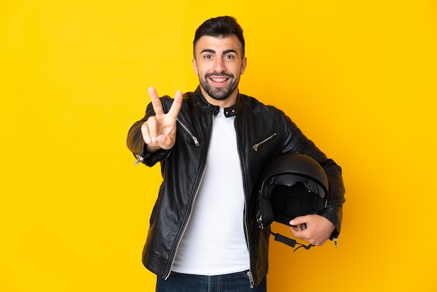 Homem caucasiano com capacete de motociclista sobre fundo amarelo isolado, sorrindo e mostrando sinal de vitória