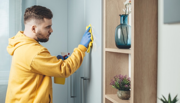 Homem caucasiano com luvas azuis limpando a casa com lenço umedecido e spray desinfetante