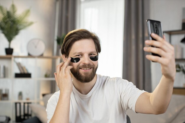 Homem caucasiano com manchas pretas de hidrogel sob os olhos, sentado em casa e falando ao telefone