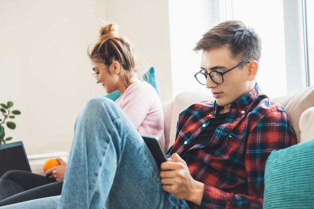 Foto homem caucasiano com óculos e sua esposa estão sentados no sofá e lendo algo em um tablet e laptop