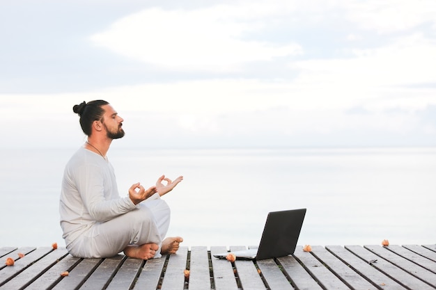 Homem caucasiano com roupas brancas meditando ioga com laptop no píer de madeira