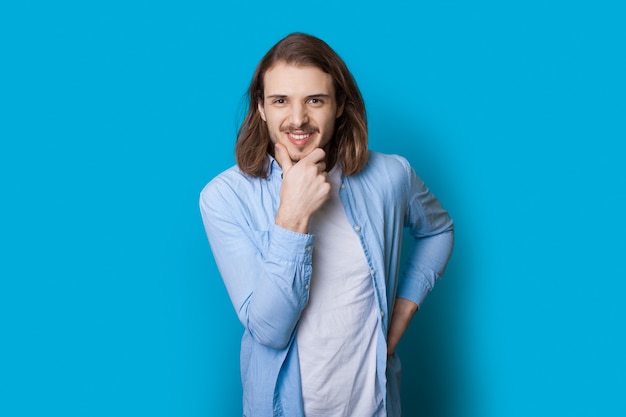 Homem caucasiano confiante com barba e cabelo comprido sorrindo com uma camisa para a câmera, tocando o queixo