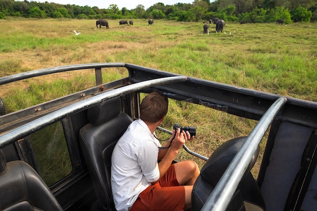 Foto homem caucasiano de camisa branca fazendo fotos de manada de elefantes durante safári no parque nacional do sri lanka