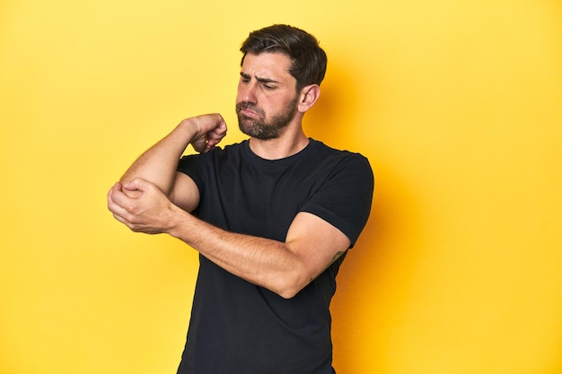 Homem caucasiano de camisa preta com fundo amarelo de estúdio massagando o cotovelo sofrendo depois de um mau movimento