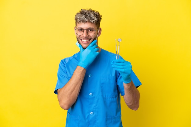 Homem caucasiano de dentista segurando ferramentas isoladas em um fundo amarelo, feliz e sorridente