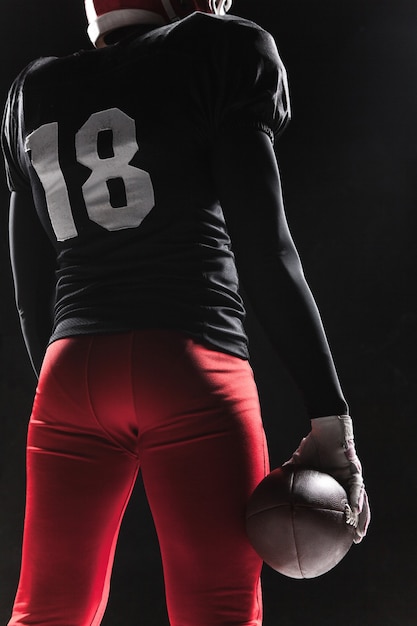 Homem caucasiano de fitness como jogador de futebol americano segurando uma bola no fundo preto
