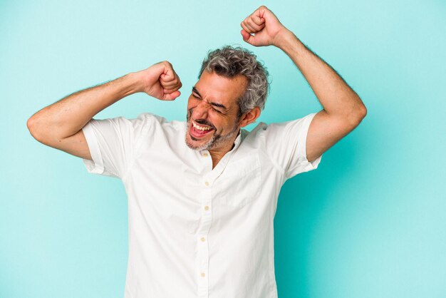 Homem caucasiano de meia-idade isolado em fundo azul comemorando um dia especial, pula e levanta os braços com energia.