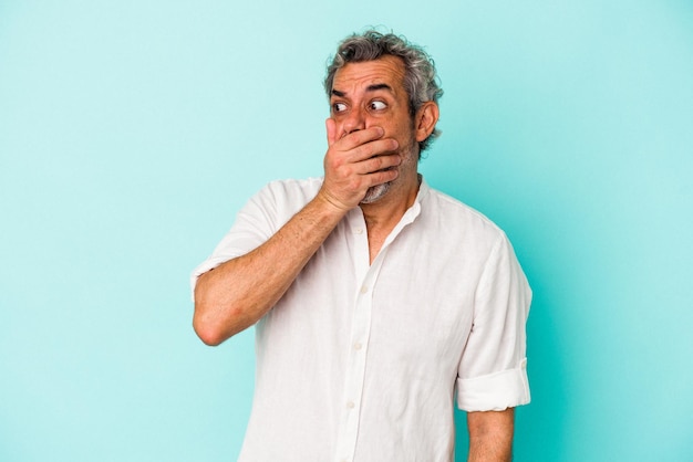 Homem caucasiano de meia-idade isolado em fundo azul pensativo olhando para um espaço de cópia cobrindo a boca com a mão.
