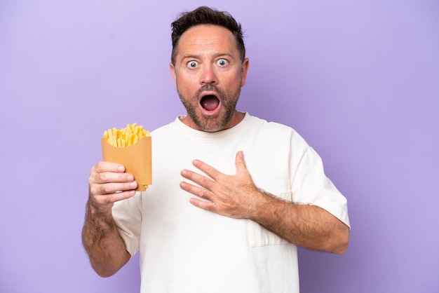 Homem caucasiano de meia-idade segurando batatas fritas isoladas em bakcground roxo surpreso e chocado ao olhar para a direita