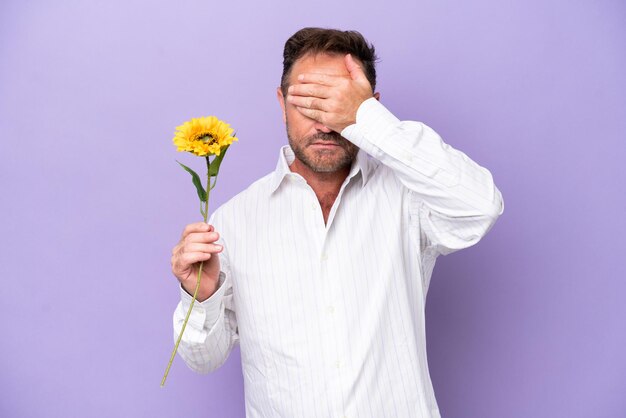 Homem caucasiano de meia-idade segurando flor de sol isolada em fundo roxo cobrindo os olhos pelas mãos Não quer ver algo