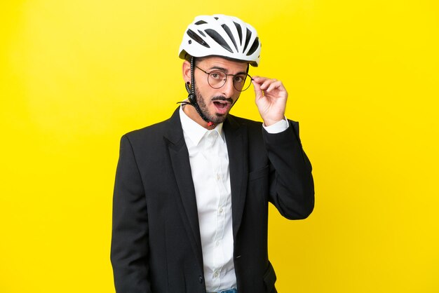 Homem caucasiano de negócios com um capacete de bicicleta isolado em fundo amarelo com óculos e surpreso