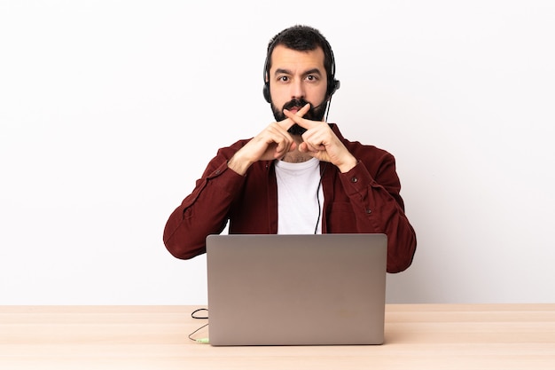 Homem caucasiano de telemarketing trabalhando com um fone de ouvido e com laptop mostrando um sinal de gesto de silêncio.