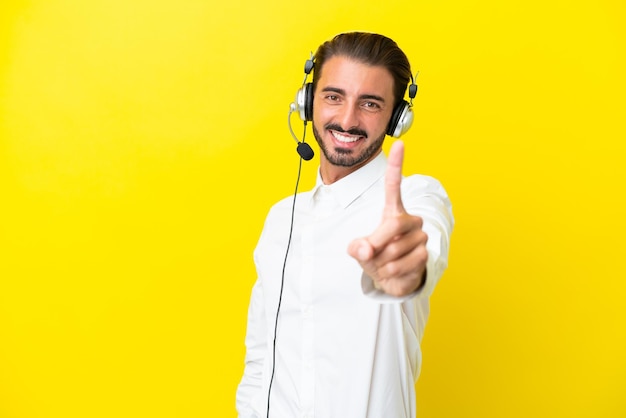 Homem caucasiano de telemarketing trabalhando com um fone de ouvido isolado em fundo amarelo mostrando e levantando um dedo
