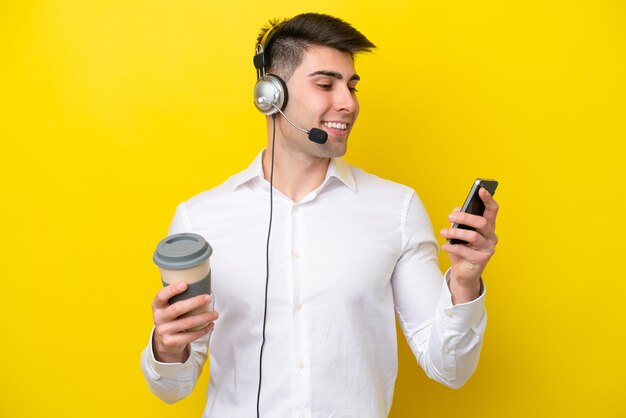 Homem caucasiano de telemarketing trabalhando com um fone de ouvido isolado em fundo amarelo segurando café para levar e um celular